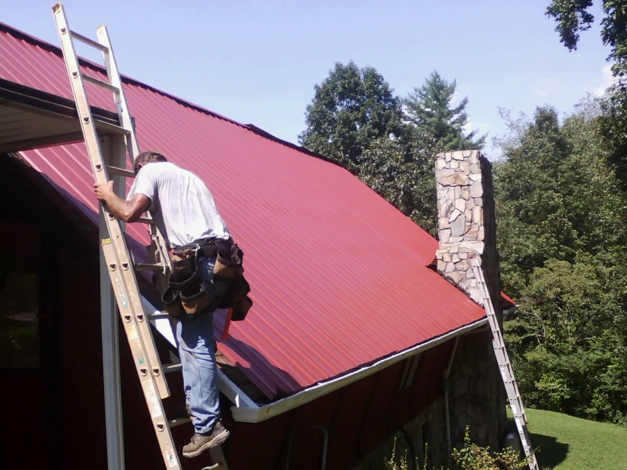 Roofer on a ladder beside a red metal roof and stone chimney — Soffit & Fascia Repair by Roofing Brothers in Farmingville